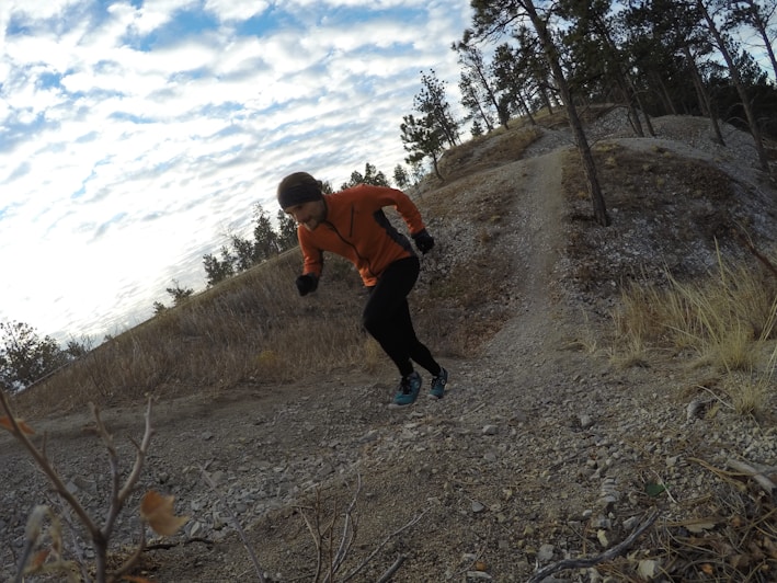 A person in an orange jacket and black pants is running uphill on a rocky trail surrounded by a natural landscape with sparse trees. The sky is partially cloudy and the terrain appears rugged with some dry grass and small bushes.
