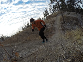 A person in an orange jacket and black pants is running uphill on a rocky trail surrounded by a natural landscape with sparse trees. The sky is partially cloudy and the terrain appears rugged with some dry grass and small bushes.