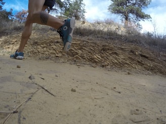 A person is running on a dirt path in a natural, rocky environment. The view is from a low angle close to the ground, capturing the motion of the runner's legs and feet. The background includes trees and a rocky hillside under a cloudy sky.