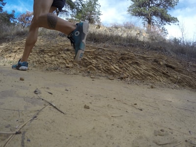 A person is running on a dirt path in a natural, rocky environment. The view is from a low angle close to the ground, capturing the motion of the runner's legs and feet. The background includes trees and a rocky hillside under a cloudy sky.