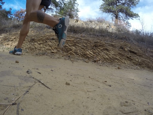 A person is running on a dirt path in a natural, rocky environment. The view is from a low angle close to the ground, capturing the motion of the runner's legs and feet. The background includes trees and a rocky hillside under a cloudy sky.