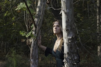 A woman stands in a forest, gazing upward with a contemplative expression. She is surrounded by tall, peeling bark trees and dense greenery, which create a natural and serene environment. The lighting casts soft shadows, highlighting the texture of the bark and the leaves.