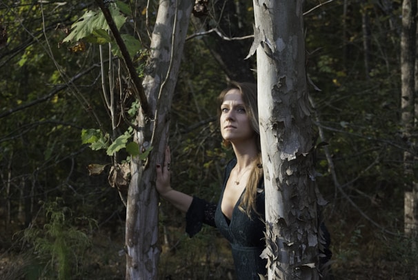 A woman stands in a forest, gazing upward with a contemplative expression. She is surrounded by tall, peeling bark trees and dense greenery, which create a natural and serene environment. The lighting casts soft shadows, highlighting the texture of the bark and the leaves.