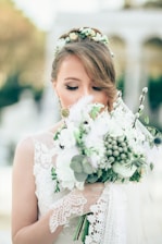 A bride with closed eyes holds a large bouquet of flowers, featuring white and green tones. Her hair is styled with a floral headpiece, and she wears a lace dress with delicate patterns. The background is softly blurred, indicating an outdoor setting with arches.