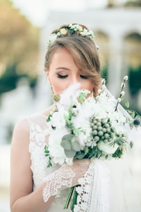 A bride with closed eyes holds a large bouquet of flowers, featuring white and green tones. Her hair is styled with a floral headpiece, and she wears a lace dress with delicate patterns. The background is softly blurred, indicating an outdoor setting with arches.