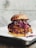 A close-up of a chicken sandwich featuring a fried chicken patty topped with red cabbage slaw, all encased in a sesame seed bun. The sandwich is placed on a white plate that rests on a dark, textured surface.