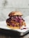 A close-up of a chicken sandwich featuring a fried chicken patty topped with red cabbage slaw, all encased in a sesame seed bun. The sandwich is placed on a white plate that rests on a dark, textured surface.