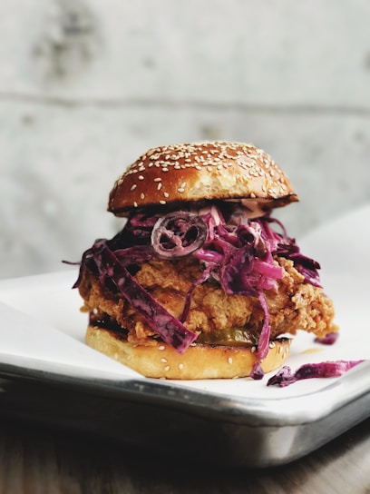 A close-up of a chicken sandwich featuring a fried chicken patty topped with red cabbage slaw, all encased in a sesame seed bun. The sandwich is placed on a white plate that rests on a dark, textured surface.