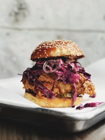 A close-up of a chicken sandwich featuring a fried chicken patty topped with red cabbage slaw, all encased in a sesame seed bun. The sandwich is placed on a white plate that rests on a dark, textured surface.