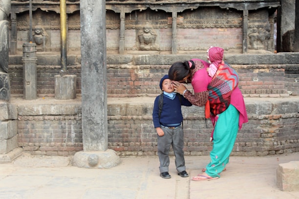 A woman in vibrant traditional clothing is bending down to interact with a child in a school uniform. They are in a rustic setting with stone and brick architecture featuring carved details. Another small child is wrapped in a colorful shawl on the woman’s back.