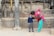 A woman in vibrant traditional clothing is bending down to interact with a child in a school uniform. They are in a rustic setting with stone and brick architecture featuring carved details. Another small child is wrapped in a colorful shawl on the woman’s back.