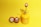 A yellow misting spray bottle with a golden nozzle is placed on a bright yellow surface. The bottle features the text 'LET it GROW' in white lettering. Next to it, two small red and white Daruma dolls are positioned on the left, characterized by their wide-eyed expressions.