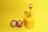 A yellow misting spray bottle with a golden nozzle is placed on a bright yellow surface. The bottle features the text 'LET it GROW' in white lettering. Next to it, two small red and white Daruma dolls are positioned on the left, characterized by their wide-eyed expressions.