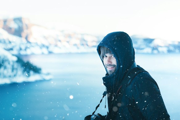 A person dressed in a dark hooded winter jacket stands in a snowy landscape. Snowflakes are visible in the air, and a distant view of snow-covered hills or mountains is blurred in the background. The person is holding a camera, giving an impression of being in a photography session in a cold, wintry environment.