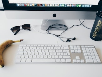 An office desk setup featuring a desktop computer with a visible Apple logo on the monitor. The desk contains a white keyboard, a banana, a pair of sunglasses, wired earphones, and a shaker bottle with the label 'aptonia'. Various app icons are displayed at the top of the computer screen.