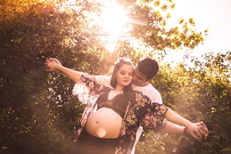 A couple is embracing in a sunlit natural setting. The woman, appearing pregnant, is wearing a floral patterned shawl and a black top, while the man stands behind her, gently holding her arms. Sunlight filters through the leaves, creating a warm, golden glow.