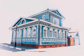 A richly decorated, traditional wooden house painted in vibrant blue. The house features intricate white carvings around the windows and along the edges, creating a striking contrast with the blue. It is a two-story building with tall windows, ornate moldings, and a steeply pitched roof. A streetlamp stands in front of the house, and the scene is brightly lit, suggesting a sunny day.
