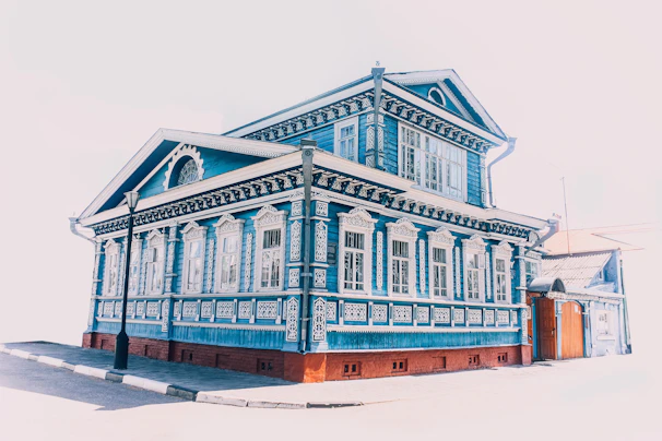 A richly decorated, traditional wooden house painted in vibrant blue. The house features intricate white carvings around the windows and along the edges, creating a striking contrast with the blue. It is a two-story building with tall windows, ornate moldings, and a steeply pitched roof. A streetlamp stands in front of the house, and the scene is brightly lit, suggesting a sunny day.