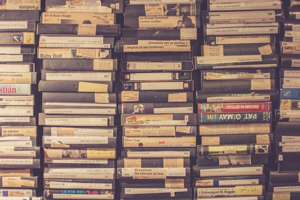 A collection of VHS tapes is neatly stacked in rows on a shelf. The spines of the tapes display various titles, many of which are written in French. The tapes vary slightly in color, with some having faded labels and others featuring colorful graphics.