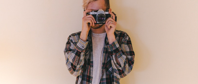 man wearing black, gray, and brown plaid collared button-up long-sleeved shirt standing while using camera