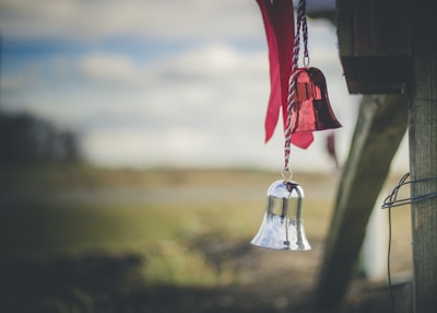 Two decorative bells hang from a wooden structure, one red and one silver, with a blurred natural background. The red bell is reflective and shiny, alongside red ribbons attached to the strings.