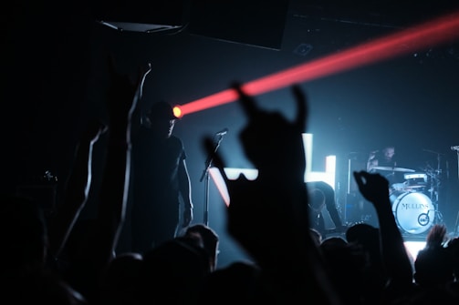 A dimly lit concert scene with a musician on stage, silhouetted against a blue backdrop, and a red laser beam cutting across the stage. The crowd's hands are raised, with some forming a rock gesture. A drummer is visible on the right playing a drum set.