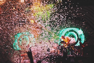 A vibrant scene at a concert with a performer playing drums surrounded by a burst of confetti. Bright green circular lights form a striking backdrop on stage, creating an energetic atmosphere.