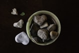 A collection of heart-shaped stones placed inside a small, round, light green dish, with additional stones scattered around it on a dark surface. The stones vary in size and texture, some being smooth while others are rough.