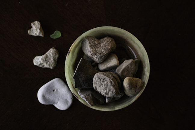 A collection of heart-shaped stones placed inside a small, round, light green dish, with additional stones scattered around it on a dark surface. The stones vary in size and texture, some being smooth while others are rough.