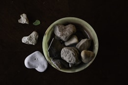 A collection of heart-shaped stones placed inside a small, round, light green dish, with additional stones scattered around it on a dark surface. The stones vary in size and texture, some being smooth while others are rough.