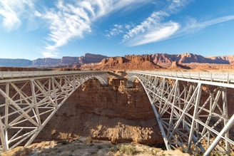 A structural steel bridge spans a deep, rocky canyon with a clear blue sky overhead and wispy clouds. The surrounding landscape is arid, featuring rugged cliffs and a vast desert.