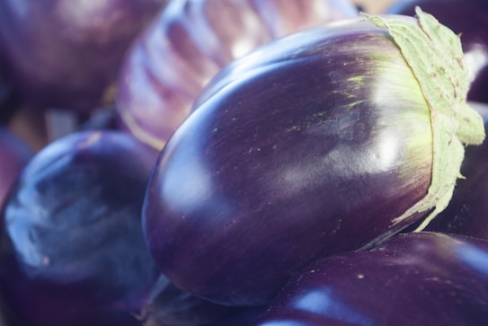 A close-up view of several glossy eggplants with vibrant purple skin and a hint of green at the stem. The focus is on the smooth texture and rich color of the eggplants, which are piled together creating an abundant and fresh appearance.