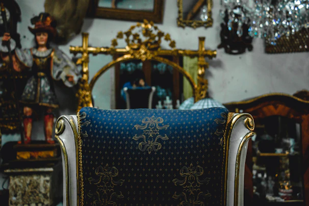 An ornate chair with a blue and gold patterned fabric is positioned in front of a large, gilded mirror. Decorative elements like a crystal chandelier and a classic statue in traditional attire are visible in the background, alongside other antique items.