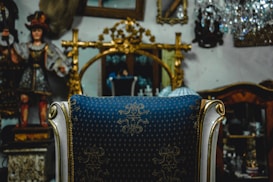 An ornate chair with a blue and gold patterned fabric is positioned in front of a large, gilded mirror. Decorative elements like a crystal chandelier and a classic statue in traditional attire are visible in the background, alongside other antique items.