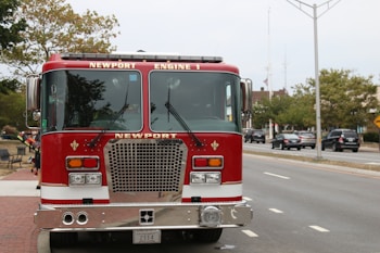 A bright red fire engine is parked by the side of a city street with clear blue skies above. The fire engine features the labels 'Newport' and 'Engine 1' on its front. Surrounding it are trees, a few parked cars off to the side, and a paved sidewalk, suggesting an urban setting.