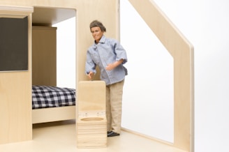 A male doll wearing a casual outfit, consisting of a light blue denim shirt and beige pants, is standing beside a bed with a black and white checkered blanket. The doll is holding onto a small wooden suitcase in a room with light wooden furniture and interior.