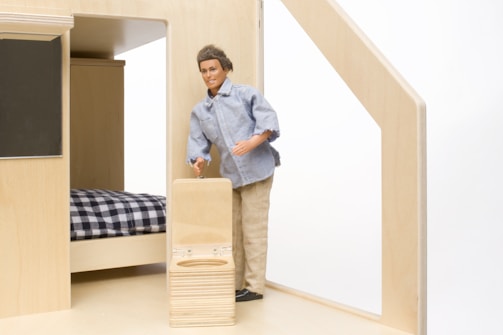 A male doll wearing a casual outfit, consisting of a light blue denim shirt and beige pants, is standing beside a bed with a black and white checkered blanket. The doll is holding onto a small wooden suitcase in a room with light wooden furniture and interior.