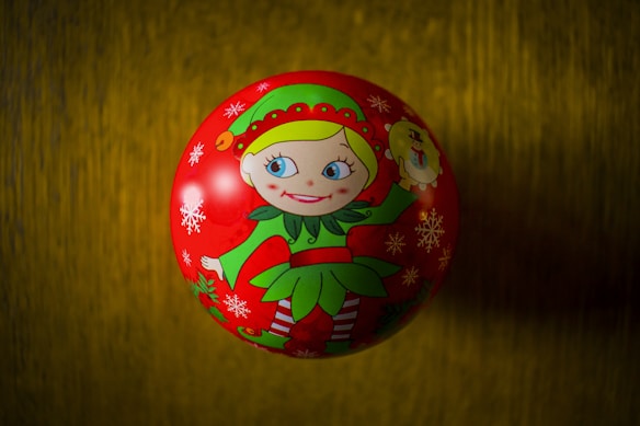 A festive Christmas ornament featuring an illustration of a cheerful elf dressed in a green and red outfit. The elf, with bright blue eyes and a big smile, is surrounded by snowflakes painted on the red surface of the ornament. The background is a muted golden color.