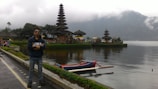 A person wearing sunglasses stands confidently near a scenic lake with a traditional, multi-tiered Balinese temple visible in the background. A small boat floats on the calm water, and misty mountains rise in the distance, creating a peaceful and tranquil atmosphere.