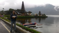 A person wearing sunglasses stands confidently near a scenic lake with a traditional, multi-tiered Balinese temple visible in the background. A small boat floats on the calm water, and misty mountains rise in the distance, creating a peaceful and tranquil atmosphere.