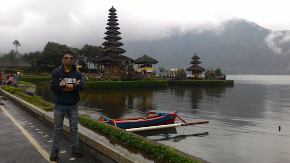 A person wearing sunglasses stands confidently near a scenic lake with a traditional, multi-tiered Balinese temple visible in the background. A small boat floats on the calm water, and misty mountains rise in the distance, creating a peaceful and tranquil atmosphere.