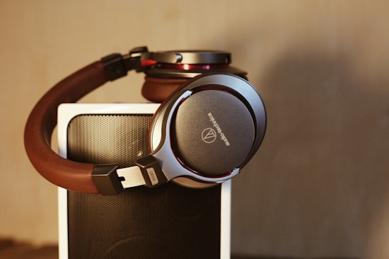Brown and silver headphones rest on top of a black speaker with a textured surface. The logo on the headphones is visible, and the lighting gives a warm tone to the scene.