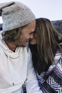 A couple is sitting closely together, smiling with their foreheads touching. The man is wearing a grey speckled beanie and a white hoodie, while the woman is wrapped in a patterned blanket or scarf with her long hair cascading down. The scene suggests warmth and intimacy.