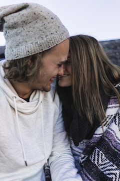A couple is sitting closely together, smiling with their foreheads touching. The man is wearing a grey speckled beanie and a white hoodie, while the woman is wrapped in a patterned blanket or scarf with her long hair cascading down. The scene suggests warmth and intimacy.