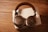 A pair of over-ear headphones with a sleek design featuring a combination of metals and dark brown accents. The headphones rest on a white box, with a wooden surface underneath, and are illuminated by warm lighting that casts soft shadows.