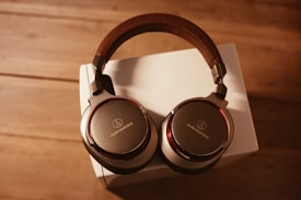 A pair of over-ear headphones with a sleek design featuring a combination of metals and dark brown accents. The headphones rest on a white box, with a wooden surface underneath, and are illuminated by warm lighting that casts soft shadows.