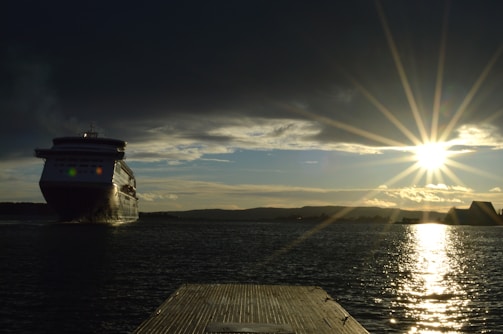 A large cruise ship is navigating through a body of water under a dramatic sky. The sun is low on the horizon, casting a bright reflection on the water, with rays of sunlight spreading across the sky.