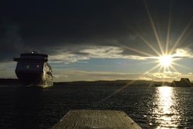 A large cruise ship is navigating through a body of water under a dramatic sky. The sun is low on the horizon, casting a bright reflection on the water, with rays of sunlight spreading across the sky.