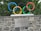 The image features five interlocking colored rings representing the Olympic symbol, mounted on a stone wall. A plaque with text is attached to the wall below the rings. Green foliage is visible in the background.