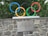 The image features five interlocking colored rings representing the Olympic symbol, mounted on a stone wall. A plaque with text is attached to the wall below the rings. Green foliage is visible in the background.
